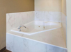 A white, corner-mounted bathtub with a chrome faucet is set in a tiled alcove with beige walls.