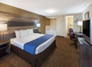 Hotel room with a king-sized bed, blue runner, two lamps, a TV, a desk chair, and a jacuzzi tub in the corner.