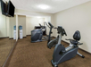 A small gym with two stationary bikes, a treadmill, a wall mirror, and a TV. Two water coolers are in the corner. The room has beige walls and brown carpet.