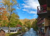 A narrow river flows beside houses and a multi-story building, surrounded by trees with autumn foliage under a clear blue sky.
