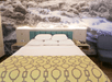 A neatly made bed with white pillows and a yellow patterned blanket is in front of a nature-themed mural featuring a river. A blue armchair is partially visible on the right.