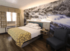 Hotel room with a bed, patterned yellow blanket, nightstand with a phone, and a gray chair. Wallpaper features a black and white nature scene of a stream with rocks.