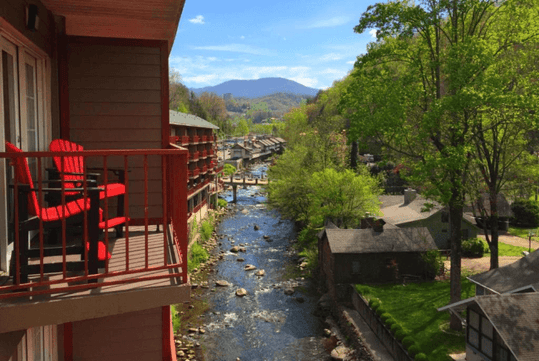 A balcony with red chairs overlooks a tranquil river surrounded by greenery, with mountains in the background under a clear blue sky.