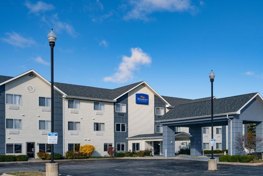 Three-story hotel building with gray roofing and beige siding under a clear blue sky. A portico extends over the entrance and two lamp posts stand in the parking area.