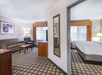 Hotel room with a living area featuring a sofa, armchair, and TV, and a separate bedroom with a double bed. Both spaces have windows with curtains and carpet flooring.