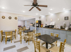 A clean dining area with several wooden tables and chairs. There is a counter with breakfast amenities, including a coffee machine and toaster. A ceiling fan is centered above.