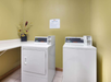 Laundry room with a white washing machine and dryer, a countertop to the left, and a decorative vase with flowers. A sign with instructions is on the wall.