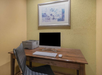 A wooden desk with a computer, keyboard, mouse, and printer. A framed abstract artwork hangs on a yellow wall above.