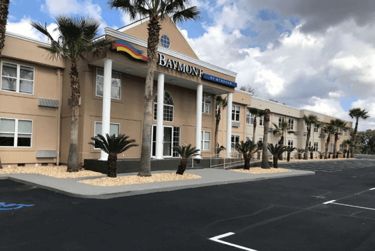 The front entrance of the Baymont by Wyndham with several palm trees in the landscaping in front of it and a blue cloudy sky over it.