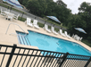 A light blue outdoor swimming pool with white lounge chairs and tables along the side and a black fence enclosing it and trees in the background.