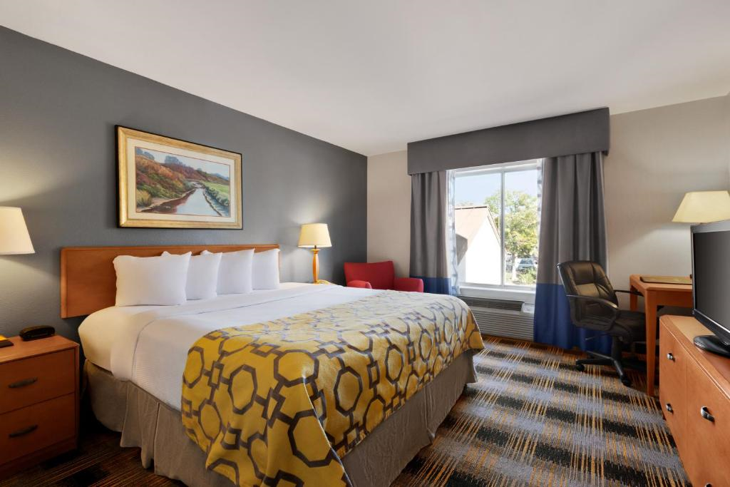 Hotel room with a double bed, patterned bedspread, side tables, lamps, armchair, desk with chair, large window, and a wall-mounted painting.