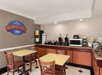 Hotel breakfast area with tables, chairs, cereal dispensers, waffle makers, microwave, beverage machine, and bread display. Sign reads "Baymont Breakfast Corner.