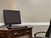 A computer monitor and keyboard on a wooden desk with a leather chair beside it. A decorative arrangement of thin stems with yellow buds is in a vase behind the monitor.