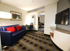 Hotel room with a blue sofa, red cushions, wall-mounted TV, desk with chair, and artwork on the wall. The room has patterned carpeting and neutral walls.