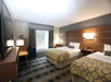 Hotel room with two double beds, a desk, chair, television, and window with curtains. Bedding includes yellow-patterned blankets. Walls are gray, with bedside lamps and wood accents.