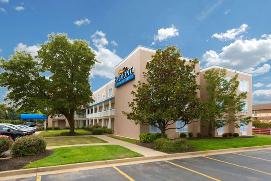 A three-story hotel building with a sign reading "Baymont" under a blue sky, with nearby trees and parked cars.