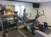 A small gym features a treadmill, stationary bike, and elliptical machine facing a mirrored wall, with a wall-mounted TV overhead.
