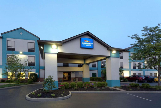 Front view of Baymont Inn & Suites building with a covered driveway, lit exterior lights, and parked cars. Trees and shrubs decorate the entrance area.