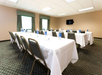 A conference room with two long tables covered in white cloths, set with water bottles, notepads, and pens, facing a wall-mounted TV screen.
