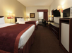 Hotel room with two double beds, a desk with a chair, wall-mounted TV, a microwave, and a painting. Beige walls and burgundy bedding. Warm lighting from lamps.