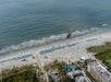 A colorful helicopter flies low over a sandy beach lined with blue umbrellas, near a hotel and pool, with waves approaching the shore.