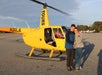 A couple stands smiling in front of a yellow tour helicopter while a pilot sits inside. The woman is showing a ring on her left hand.