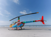 A colorful helicopter with the registration N442BD is parked on a sandy beach with the ocean and blue sky in the background.
