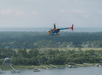 A colorful helicopter flies over a waterfront area with docks, houses, trees, and a view of the distant ocean.