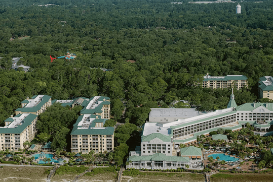 Aerial view of a resort complex with multiple buildings, pools, and landscaped grounds surrounded by dense forest; a helicopter is flying above the trees.