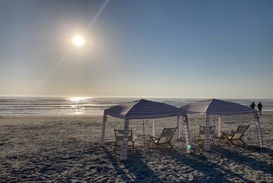 Beach scene at sunset with three striped cabanas and deck chairs facing the ocean. Two people stand near the water's edge.
