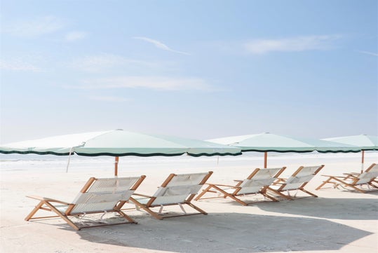 Wooden lounge chairs and white umbrellas line a sunny beach with clear skies and calm waves.