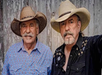 Two older men wearing cowboy hats stand in front of a wooden wall. One wears a blue patterned shirt, the other a black shirt with embroidery.