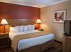 Hotel room with a large bed, four pillows, two bedside lamps, a painting on the wall, and a television on a dresser.