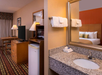Hotel room featuring a bed, side table, TV on a dresser, desk with a lamp, mini fridge, and a bathroom vanity with towels and a mirror.