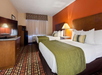 Hotel room with two double beds, a TV on a dresser, a small fridge, and an orange accent wall. A window with curtains and a patterned carpet complete the decor.