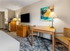 Hotel room interior with a bed, wooden table and chairs, a dresser with a TV, two lamps, and a framed artwork on a light-colored wall.