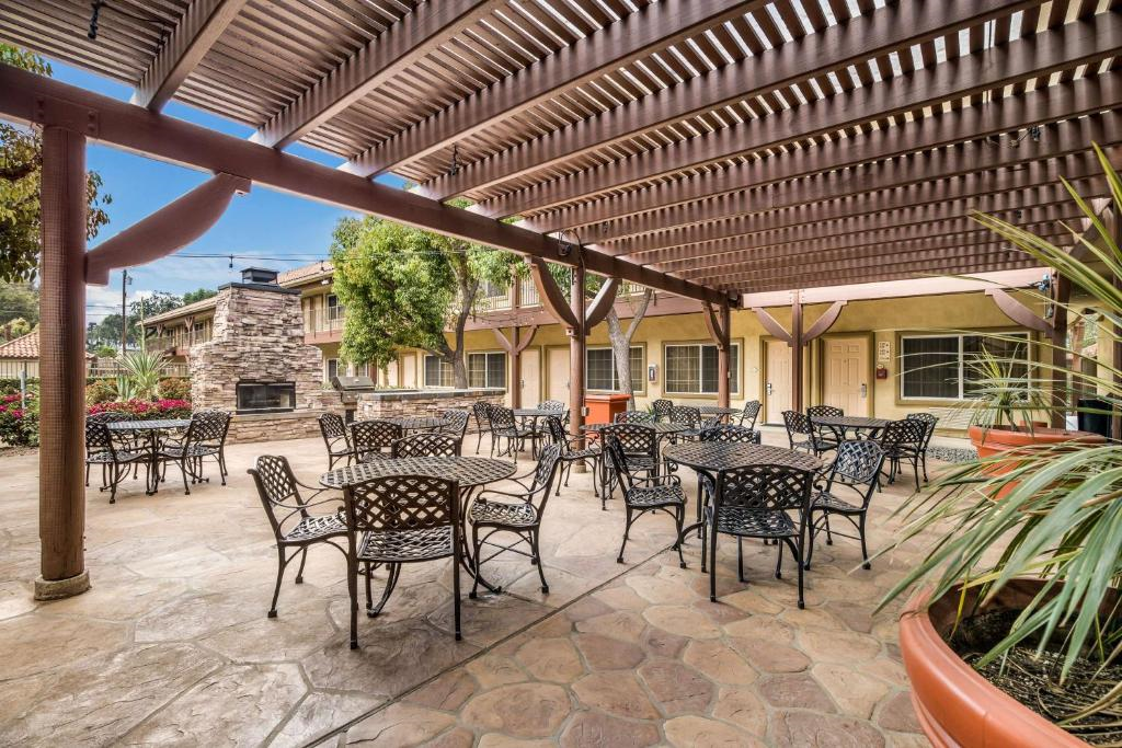 Outdoor patio with metal tables and chairs under a wooden pergola. Stone flooring and a stone fireplace are in the background. Lush greenery is visible around the area.