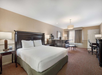 Hotel room with a queen-sized bed, wooden furniture, two lamps, a desk, a window with curtains, and a lounge chair. Neutral color palette with beige walls and carpet.