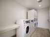 A laundry room with four front-loading washing machines, two stacked on the right, and a folding table on the left. The floor is tiled, and the room is lit by an overhead light.