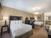 Hotel room with two double beds, bedside tables with lamps, a small seating area, and a coffee station. Neutral tones and a wall-mounted framed picture complete the decor.