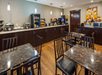 Hotel breakfast area with dark wood tables and chairs. Counter along the wall has food items, beverages, and kitchen appliances. Wall art and overhead lights are visible.