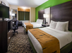 Hotel room with two double beds, green accent wall, patterned carpet, desk with chair, flat-screen TV, and a window with curtains.