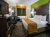 Hotel room with a king-size bed, dark wood furniture, television, desk, and chair. Green accent wall and bed runner, and a window with curtain near an air conditioning unit.