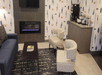 A modern hotel lobby with a sofa, two chairs, a coffee table, a wall-mounted TV, an electric fireplace, and a coffee station. The wallpaper features a blue and yellow abstract pattern.