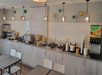 Breakfast buffet area with self-serve food stations, beverage dispensers, and seating. Several lights hang from the ceiling, and colorful wall decorations are displayed above the counter.