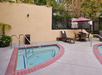 Outdoor hotel area with two small jacuzzi pools, a lounge chair, a table with four chairs under a red umbrella, and a surrounding fence and greenery.