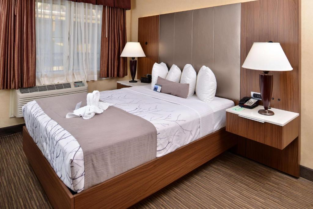 Hotel room with a neatly made bed, three pillows, folded towel swan decoration, and bedside lamps. A window with curtains is on the left and a telephone on the right nightstand.