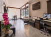 Hotel lobby with tiled floor, seating, a wall-mounted TV, desk with computer, and a plant with pink flowers. Large windows provide natural light.