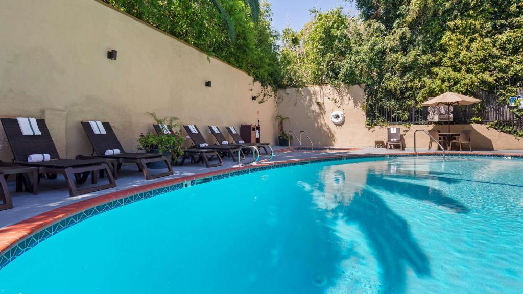 An outdoor pool with clear blue water is surrounded by lounge chairs with towels. A wall with greenery is in the background, and there's a table with an umbrella on the deck.