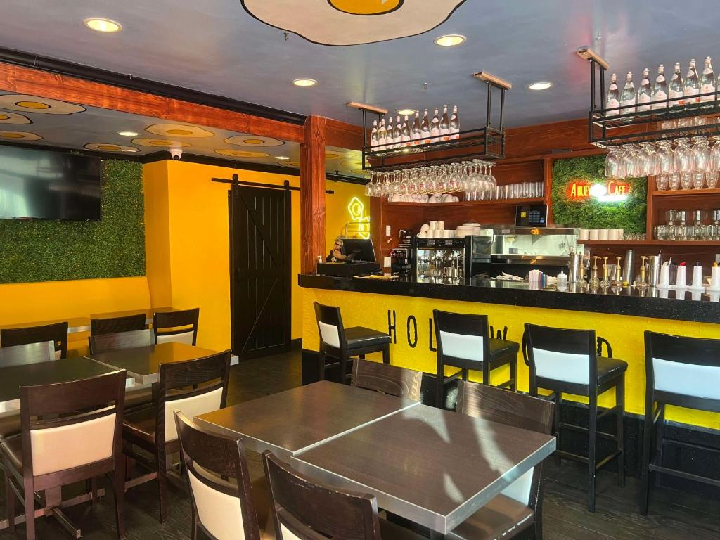 A modern cafe interior with yellow accents, wooden tables, chairs, and a counter with a coffee machine. Glassware is hung above the bar area, and a neon coffee sign is on the wall.
